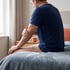 A man in a blue shirt sits on the edge of a bed and holds his elbow.