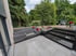 Moderne Terrasse im Bau mit hellgrauen Steinplatten, schwarzen Stufen und betonierten Hochbeeten vor grünem Garten.