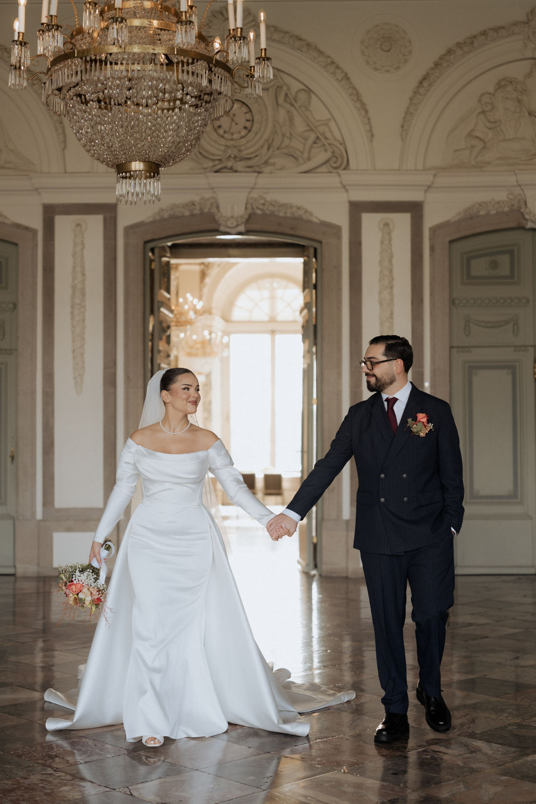 Brautpaar geht Hand in Hand unter dem Kronleuchter durch Schloss Benrath in Düsseldorf.
