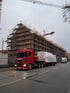 Roter Scania-LKW mit weißer Ladung auf der Straße vor einem eingerüsteten Gebäude mit Kränen.
