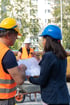 Bauarbeiter in Warnweste und Ingenieurin mit Helm betrachten gemeinsam einen Bauplan auf einer Baustelle.