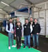 Sechs lächelnde SKP-Mitarbeiter in schwarzen Westen stehen vor einer Industrieanlage auf einer Messe mit grünem Boden.