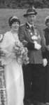 Eine lächelnde Frau in einem Hochzeitskleid mit Diadem und ein Mann in einer Uniform mit Hut, die einen Blumenstrauß hält.