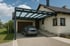 Graues Auto unter einem modernen Carport mit Glasdach neben einem hellen Haus mit Garage.
