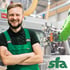 Lächelnder Mann mit Bart in grüner Arbeitskleidung vor Maschinen, unten rechts "sfa"-Logo.