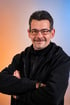 Ein lächelnder Mann mit Brille und Schnurrbart, die Arme verschränkt, trägt einen schwarzen Kapuzenpullover.