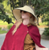 Blonde woman in a red cape, sun hat, and sunglasses looking to the right.