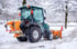 Grüner Traktor mit Schneepflug und Streuer räumt Schnee von einem Weg.