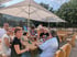 Eine Gruppe von Menschen sitzt an einem Holztisch im Biergarten unter weißen Veltins-Sonnenschirmen im Freien.