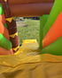 Aufsicht auf eine bunte Hüpfburg mit grünen, orangefarbenen und braunen Elementen, Gras und Garten im Hintergrund.