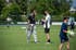 Zwei Fußballtrainer in Sportkleidung unterhalten sich auf einem grünen Feld, während Kinder im Hintergrund spielen.