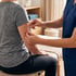 A healthcare worker in blue scrubs applies a clear nicotine or hormone patch to a patient's upper arm.