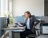 A woman in a gray blazer sits at an office desk holding papers and touching her lower back with a pained expression.