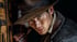A man in a weathered fedora looks intently at the viewer, his face smudged with dirt.