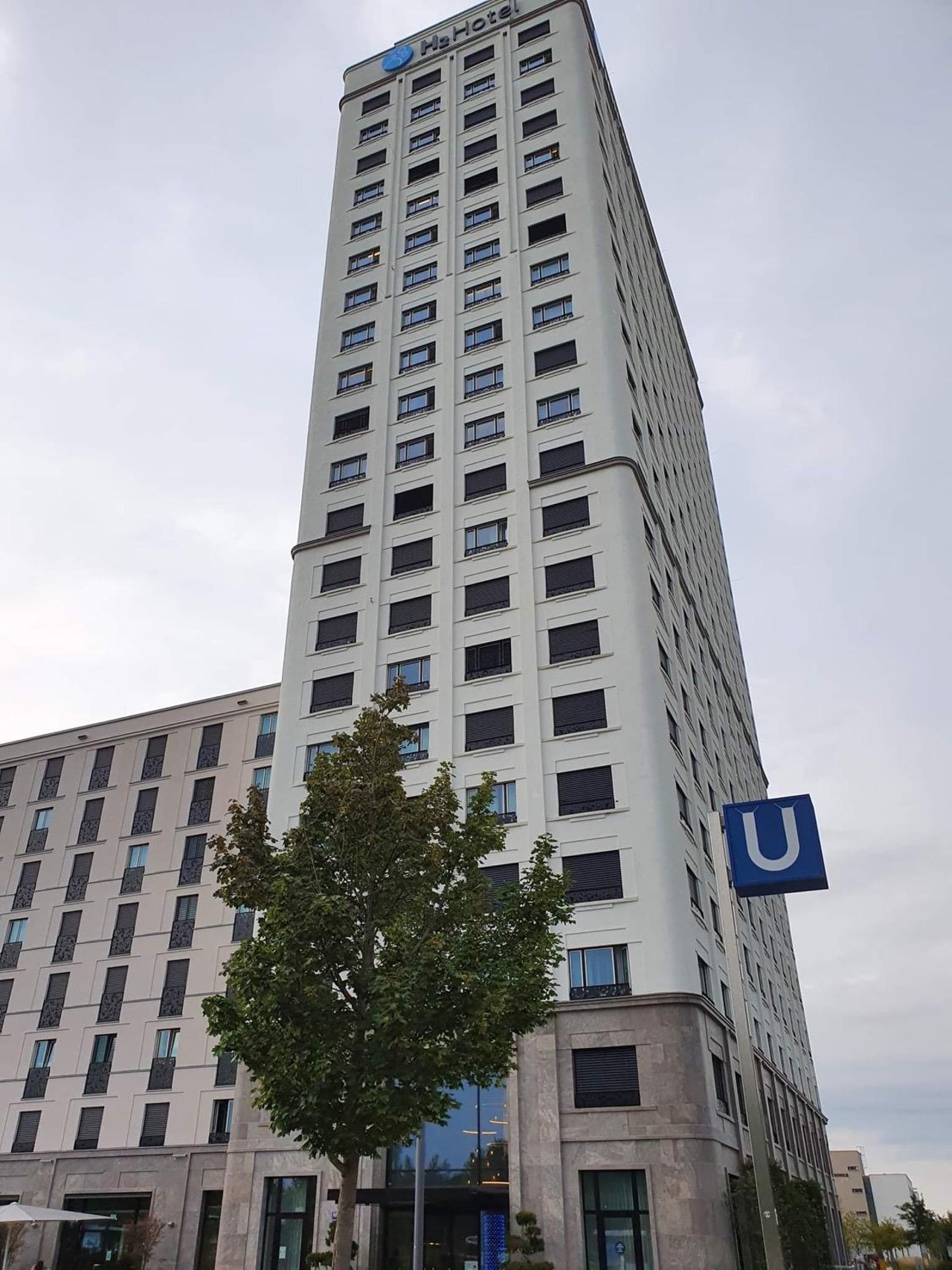Hohes weißes Gebäude mit Hs-Hotel-Logo, einem Baum und einem blauen U-Bahn-Schild.