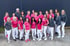 Eine Gruppe von 19 Personen, meist Frauen, in pinkfarbenen Poloshirts und weißen Hosen vor einer grauen Wand.