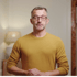 Mann mit grauem Haar, Brille und gelbem Pullover blickt ernst direkt in die Kamera.