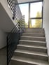 Treppe mit grauen Fliesen, schwarzem Metallgeländer und einem großen Fenster mit Blick auf Bäume.