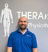 Lachender Physiotherapeut in blauem Poloshirt vor einer grauen Wand mit "THERAnove" Logo.