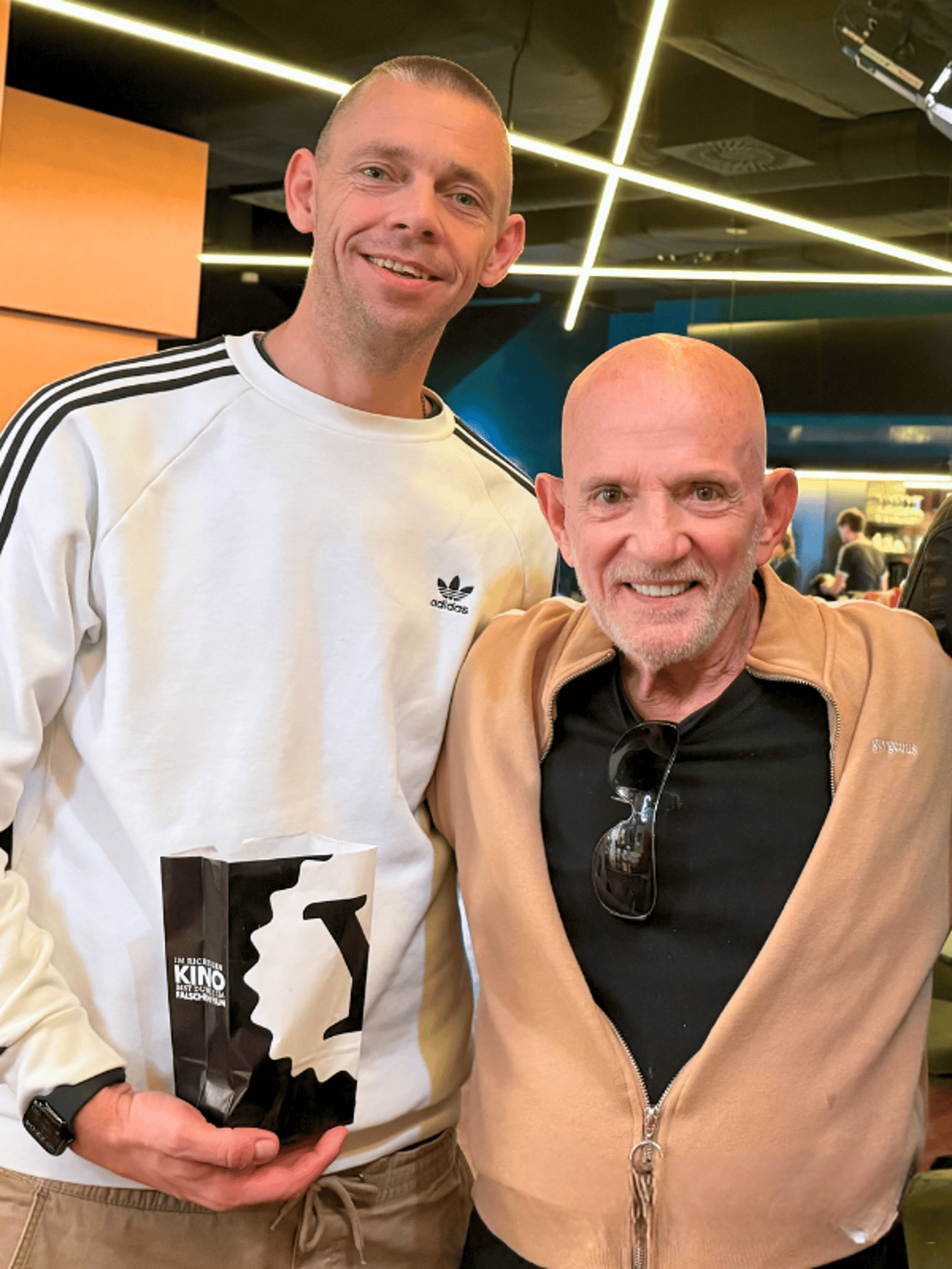 Two men smile at the camera, one holding a popcorn bag reading KINO.