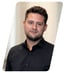 Ein lächelnder Mann mit dunklen, kurzen Haaren und Bart trägt ein schwarzes Hemd vor einem hellen Hintergrund.