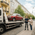 Ein Abschleppwagen mit einem roten Auto auf der Ladefläche steht auf der Straße; ein Mann und eine Frau stehen daneben.
