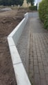 L-Steine aus Beton bilden eine Stützmauer zwischen einem gepflasterten Gehweg mit Schotterstreifen und einer Erdfläche.