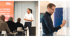 Ein Mann in weißem Hemd spricht mit zwei jüngeren Männern; ein Mann in schwarzem Hemd schreibt auf eine Tafel.
