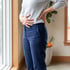 A person in a grey sweater and blue trousers stands by a window, holding their lower back and abdomen.