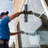 Mann in blauem Hemd und Kappe reinigt mit Fensterwischer ein großes Glasfenster an einem Gebäude.
