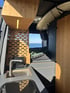 Campervan-Innenraum mit Sicht auf das Meer, gemütlichem Bett unter einem Surfbrett, durchdachter Trennwand, LED-Beleuchtung und moderner Edelstahlspüle – perfekter Ausbau für Abenteurer.
