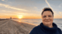 Lächelnde Person im blauen Rollkragenpullover am Strand vor einem Sonnenuntergang über dem Meer.