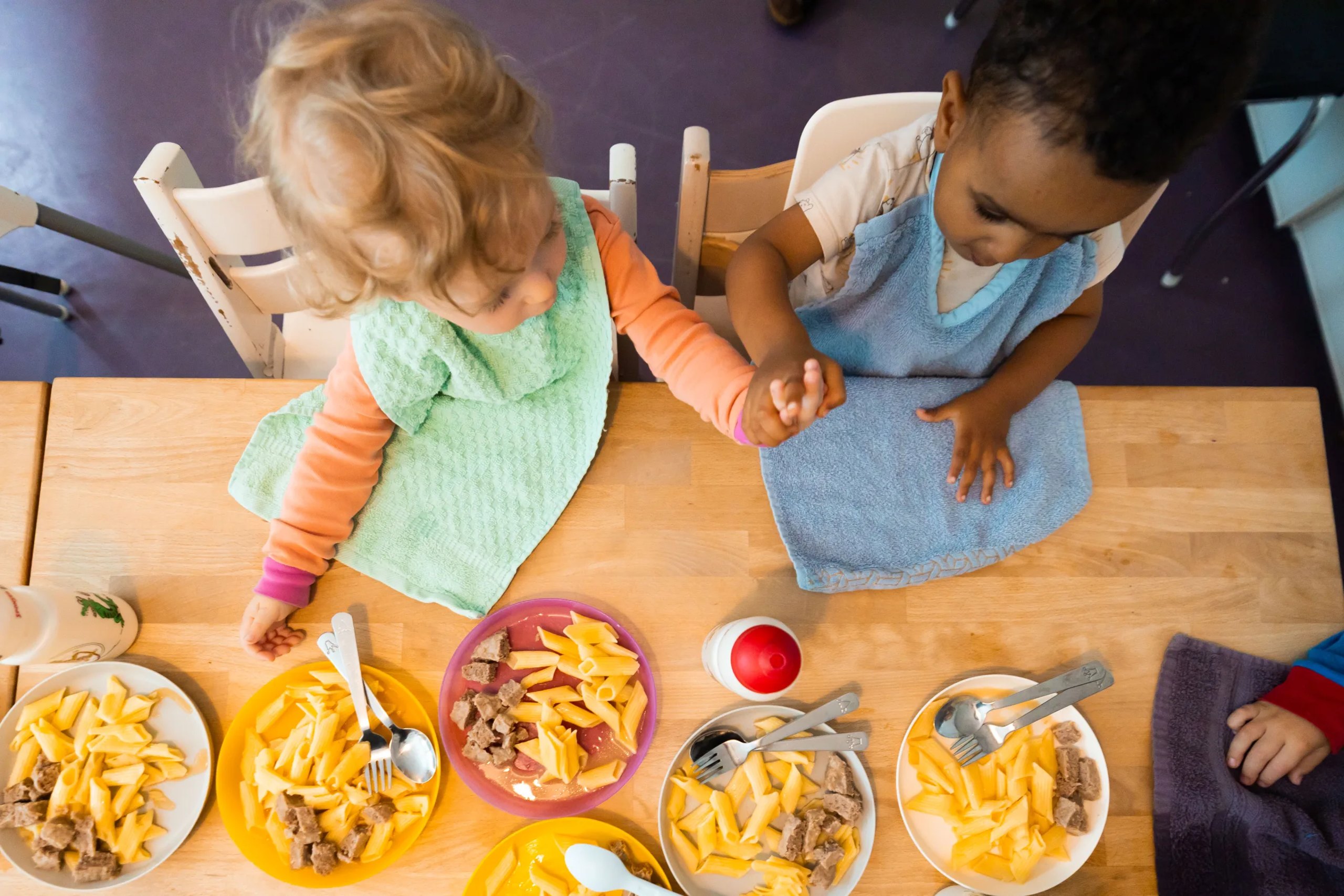 Zwei Kinder halten sich am Mittagstisch über den Tellern die Hände.