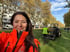 Frau in orangefarbener Warnjacke macht ein Selfie im Park, während im Hintergrund ein Traktor auf der Wiese arbeitet.