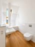 Modernes Badezimmer mit Dachschräge, weißem Waschbecken, Wand-WC, Badewanne, Holzfußboden und hellem Dachfenster.