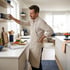 A man in an apron winces and clutches his lower back while reaching for a bowl on a high shelf in a modern kitchen.