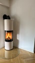 Moderner, zylindrischer Kaminofen mit brennendem Holzfeuer hinter Glas auf einer runden Glasplatte vor weißer Wand.