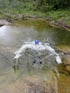 Person mit blauem Helm im Wasser, das spritzt, mit bewaldeten Ufern im Hintergrund.