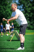 Ein männlicher Trainer in weißem T-Shirt, schwarzen Shorts und neongrüner Brille bückt sich auf einem Fußballfeld.