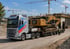 Ein Volvo-Schwertransport-LKW transportiert einen gelben CAT-Kettenbagger auf einer Baustelle mit Kränen im Hintergrund.