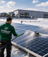 Ein Mann in grüner "PUTZPALAST REINIGUNGSSERVICE" Uniform reinigt Solarmodule auf einem Dach mit einem langen Sprühgerät.