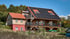 Großes Haus mit Holzfassade, rotem Dach und Solarpaneelen; rotes Erdgeschoss, Balkon mit Pflanzen.