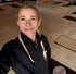 Lächelnde Frau mit blonden Zöpfen in schwarzer Sportjacke steht in einem Yoga-Studio mit ausgelegten Matten.