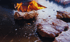 Mehrere Fleischstücke braten auf einer großen, runden Grillplatte mit Flammen im Hintergrund.