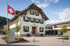Schweizer Gemeindehaus mit grünen Fensterläden, wehender Landesflagge und modernem Vorplatz unter blauem Himmel.