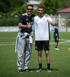 Zwei lächelnde Männer in Sportkleidung geben auf einem grünen Fußballfeld die Daumen hoch.
