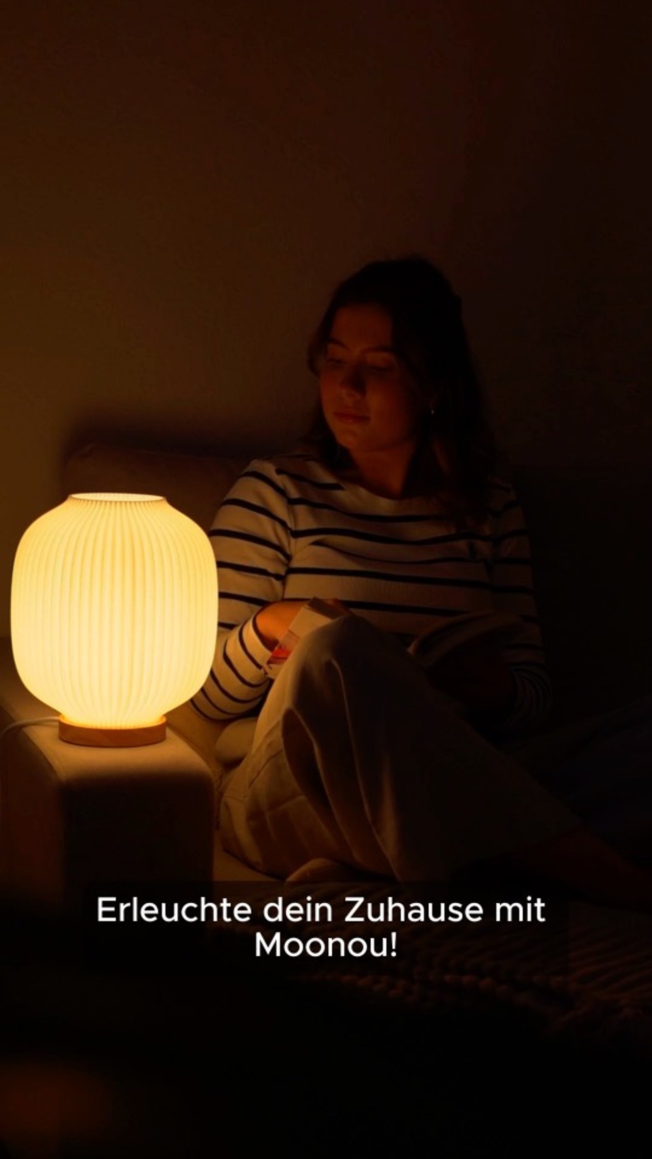 Standbild aus einem Moonou Werbevideo: Frau liest abends auf dem Sofa neben einer leuchtenden Moonou Tischlampe, Untertitel Erleuchte dein Zuhause mit Moonou