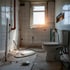 Unfertiges Badezimmer mit weißen Fliesen, sichtbaren Rohren, Kabeln und einer Toilette; Sonnenlicht scheint durch das Fenster.