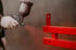 Hand in weißem Handschuh sprüht roten Lack auf ein rotes Metallteil mit einer Spritzpistole.