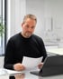 Ein Mann mit Bart im schwarzen Pullover betrachtet ein Dokument vor einem Laptop in einem hellen Büro.